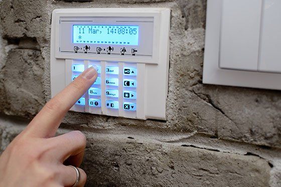 Dito di una persona che preme i pulsanti su una tastiera bianca di un sistema di sicurezza montata su un muro di mattoni. Lo schermo mostra data e ora.