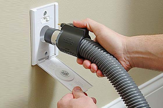 Inserimento manuale di un tubo flessibile per aspirapolvere in una presa per aspirapolvere montata a parete, piastra bianca, tubo flessibile grigio, parete marrone chiaro.