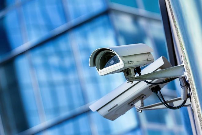 Due telecamere di sicurezza bianche montate sull'esterno di un edificio con una facciata in vetro blu sfocato sullo sfondo.