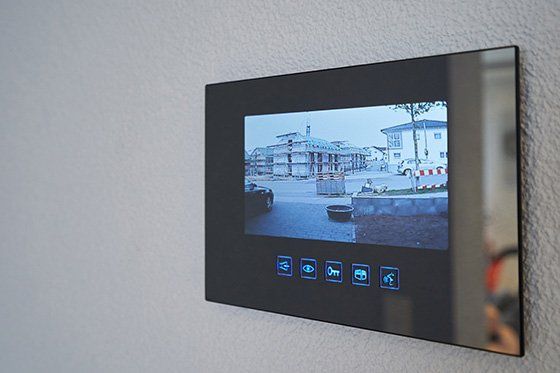 Monitor videocitofonico nero montato su una parete bianca, che mostra una vista esterna di un edificio in costruzione.