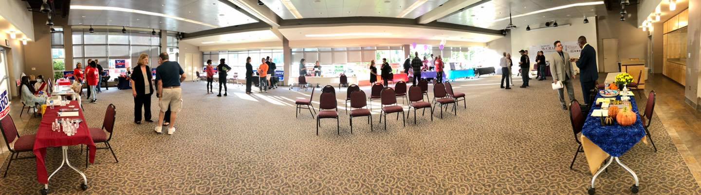 A wide shot of an event in a large hall. Tables are set up with people mingling and a few rows of chairs in the center.