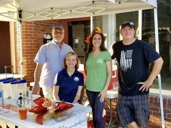 Four people stand behind a table under a white canopy, likely at an outdoor event. The woman in green smiles; the man on the right wears an American flag shirt.