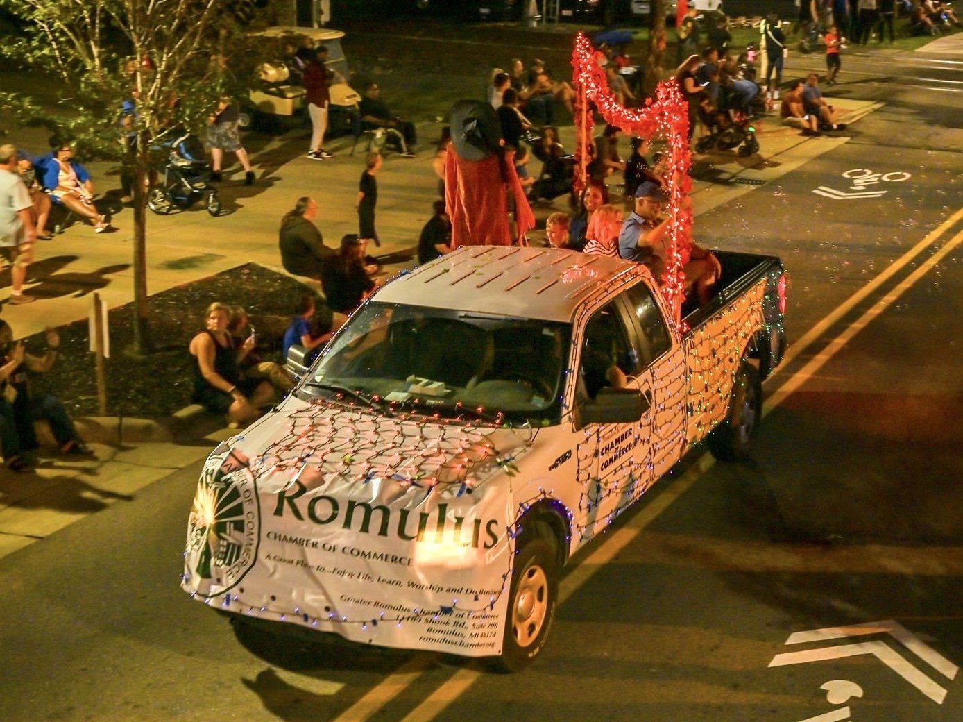 A decorated white truck in a nighttime parade with people riding in the bed, lit by lights. Spectators line the street.