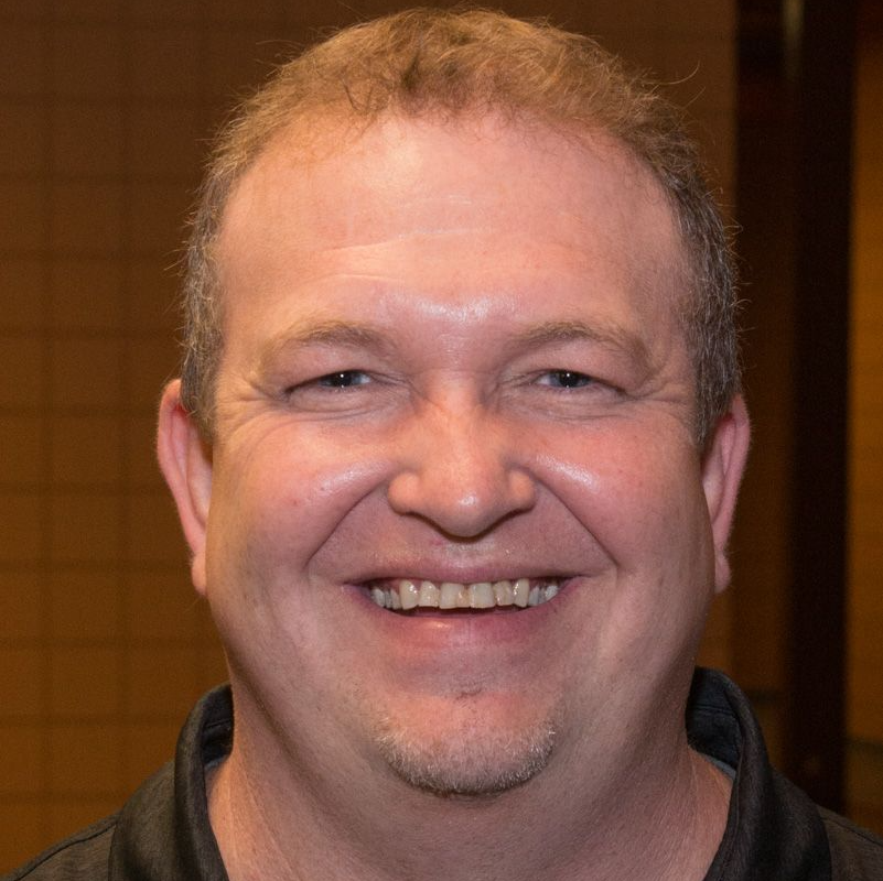 Smiling man with short, light brown hair and a goatee, wearing a black collared shirt, indoors.