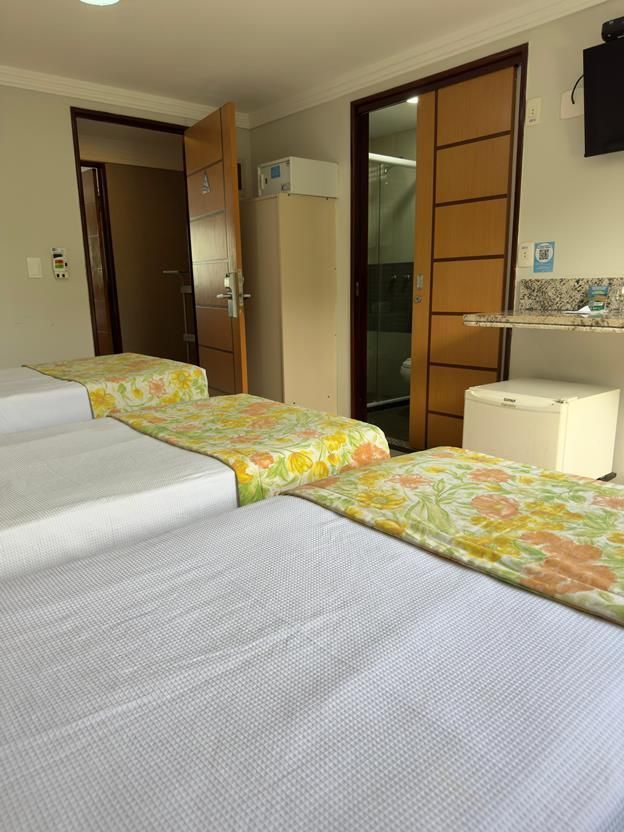 Um quarto de hotel com três camas com colchas amarelas estampadas com flores, duas portas de madeira e um pequeno frigobar branco.