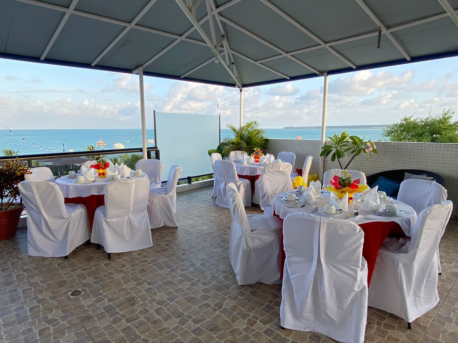 Uma área de jantar externa e coberta com vista para o oceano, com mesas redondas cobertas com toalhas de mesa e cadeiras brancas.