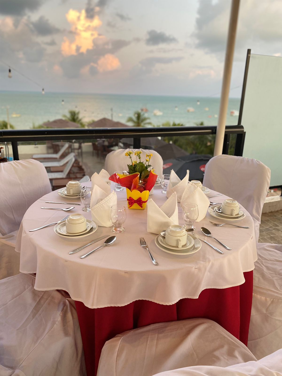 Uma mesa redonda posta para quatro pessoas, com toalhas de linho branco e um pequeno vaso amarelo, com vista para um mar calmo ao pôr do sol.