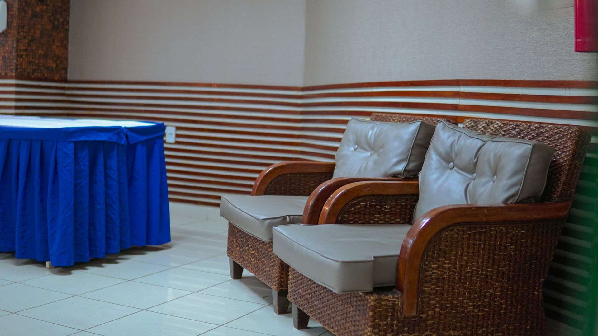 Duas poltronas de vime com almofadas cinza-claro ficam ao lado de uma mesa coberta com uma toalha de mesa azul em uma sala com paredes listradas.