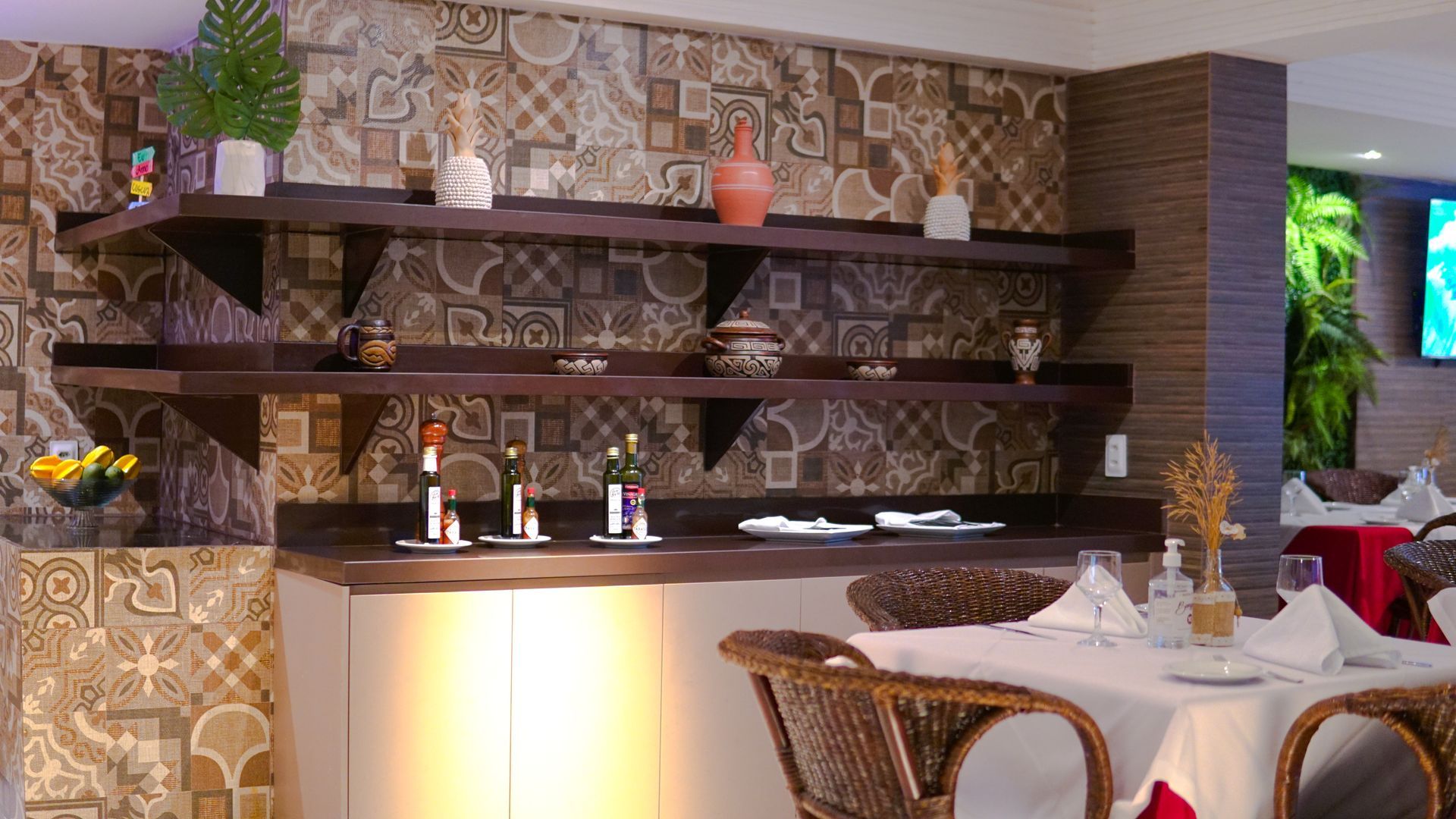 Interior de restaurante com paredes revestidas de azulejos, prateleiras de madeira escura com objetos decorativos e mesas com cadeiras de vime.