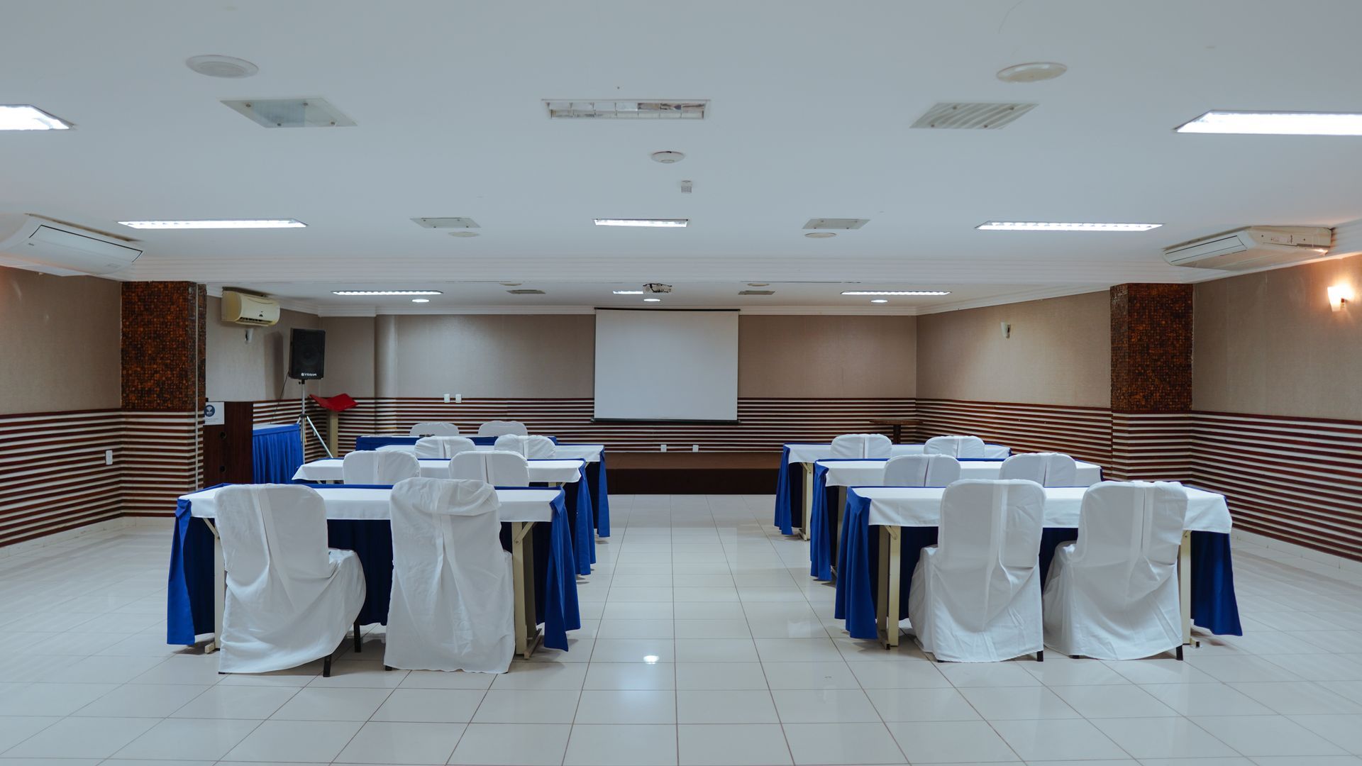 Uma sala de reuniões com fileiras de mesas com toalhas de mesa brancas, saias azuis e cadeiras brancas voltadas para uma tela de projeção.