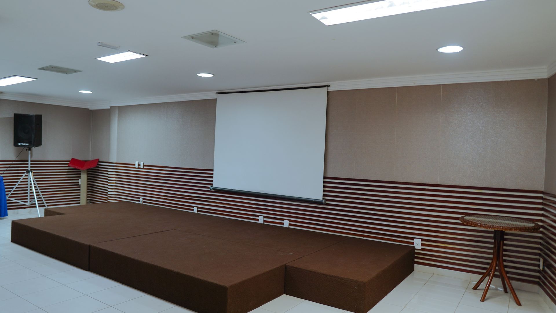 Um palco elevado com uma tela de projeção, uma caixa de som em um tripé e uma pequena mesa lateral em uma sala com paredes listradas.