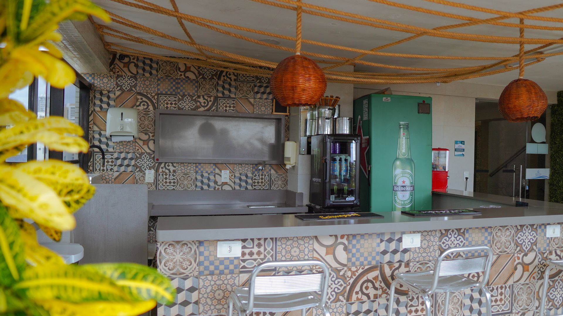Um bar ao ar livre com balcões e paredes revestidos de azulejos, luminárias suspensas de vime e duas cadeiras sob um teto de palha.