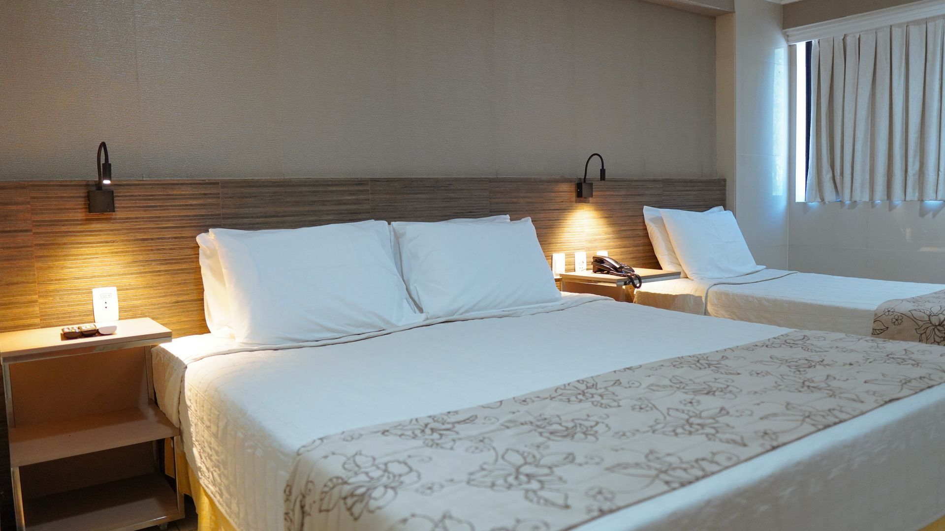 Um quarto de hotel luminoso com uma cama grande em primeiro plano, uma cama de solteiro ao lado, mesas de cabeceira e uma janela com persianas.