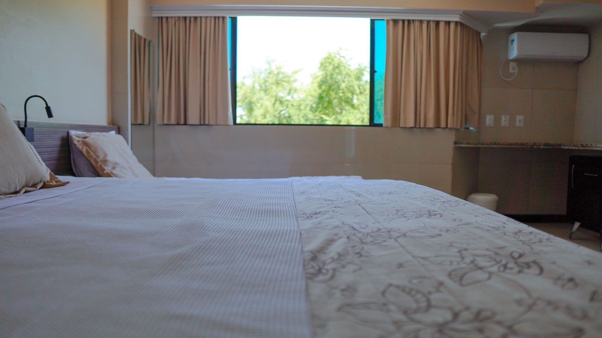 Um quarto de hotel arrumado com uma cama grande, roupa de cama com estampas claras e uma janela com vista para árvores verdes.