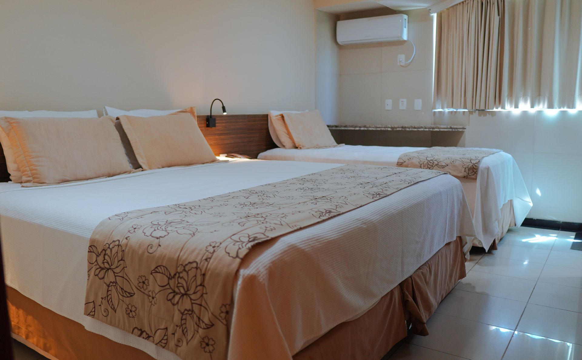 Um quarto de hotel com duas camas de solteiro com roupa de cama bege e caminhos de mesa florais, ao lado de uma janela com cortinas claras.