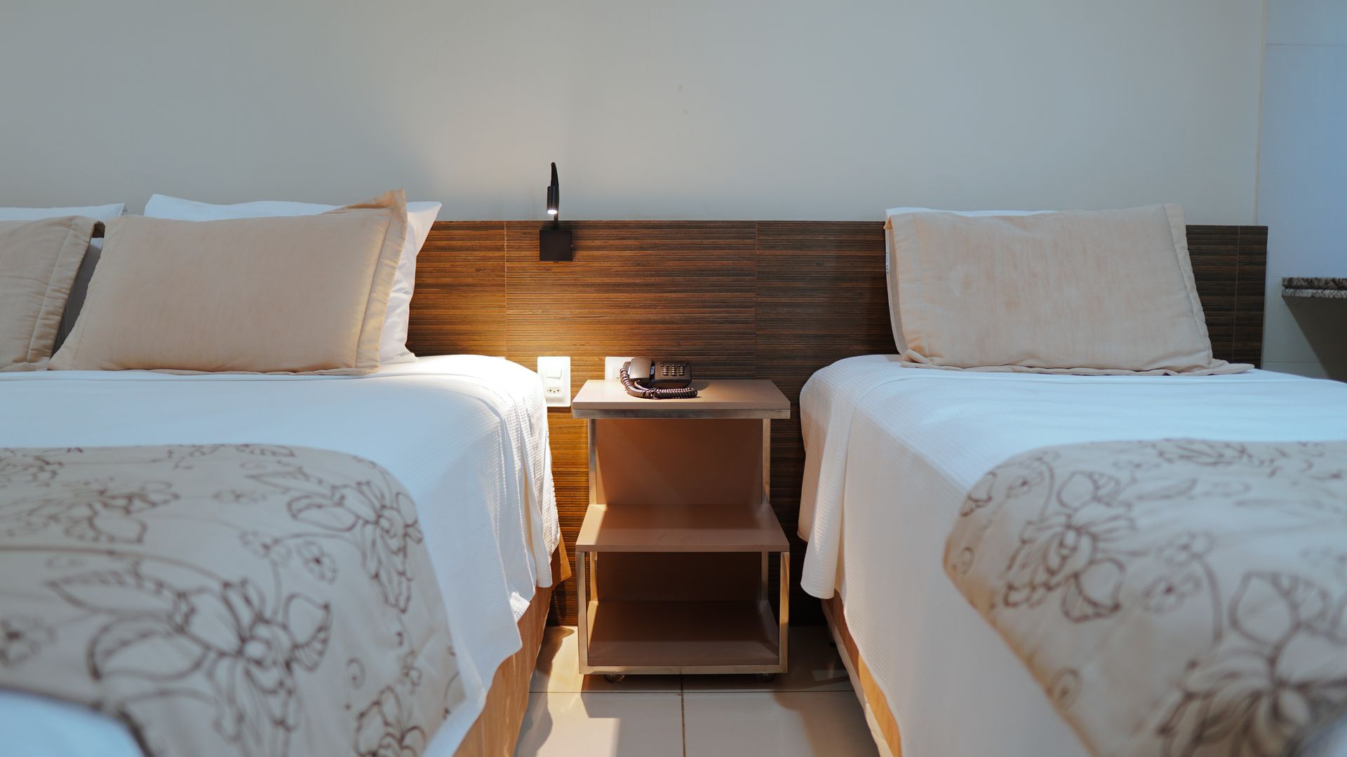 Duas camas de solteiro com roupa de cama e travesseiros em tons de bege claro, separadas por uma pequena mesa de cabeceira de madeira com dois níveis.