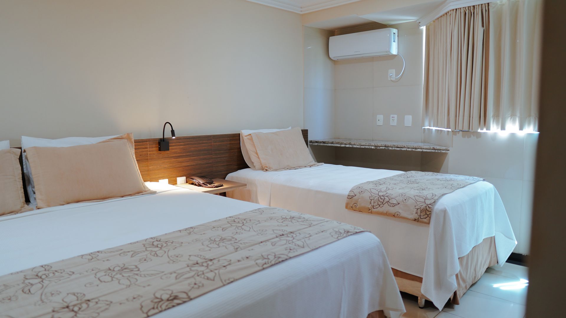 Um quarto de hotel com duas camas de solteiro, lençóis brancos, peseiras e travesseiros bege, em uma cabeceira de madeira escura.