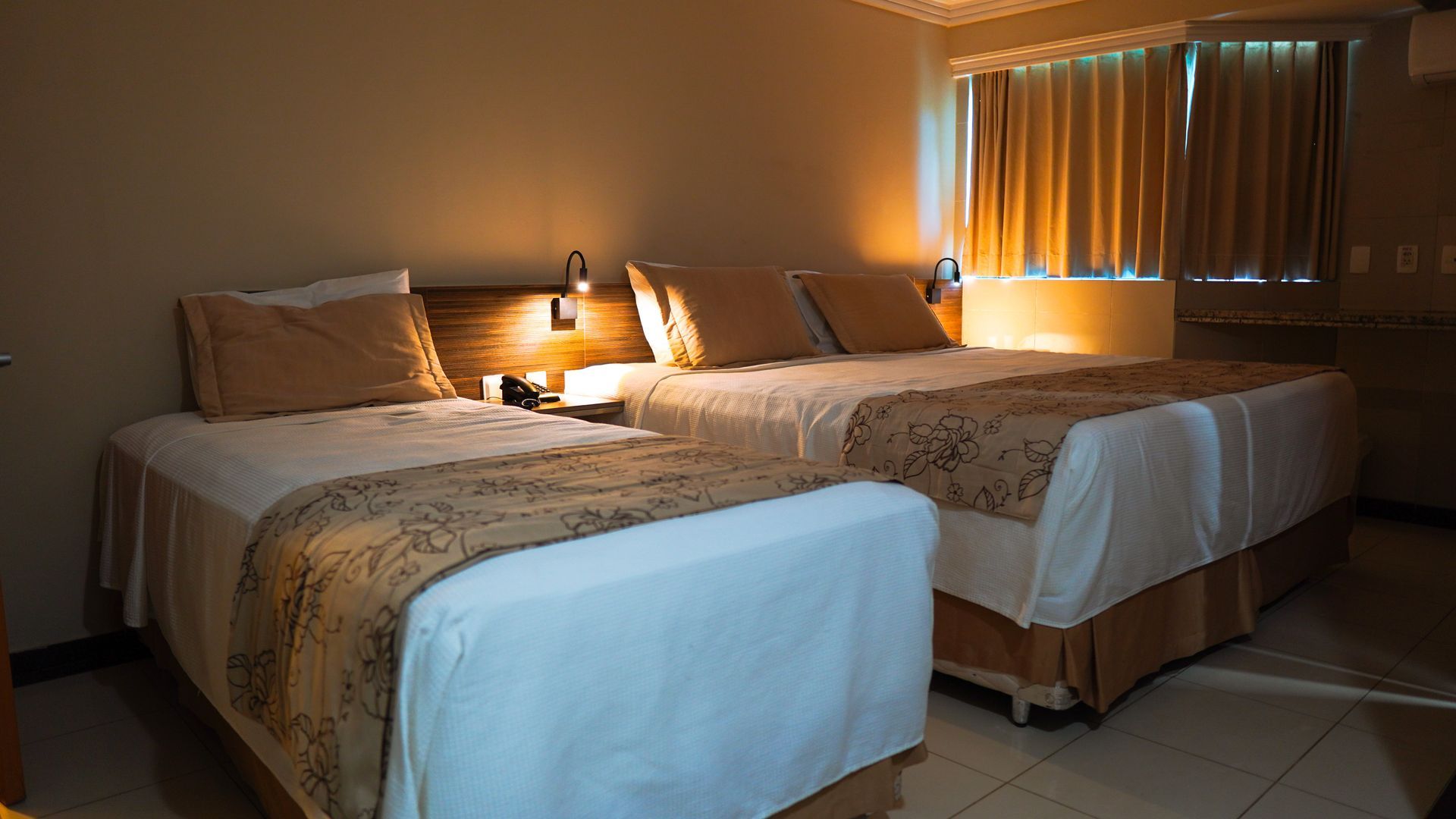Um quarto de hotel com duas camas, lençóis brancos, passadeiras marrons estampadas, travesseiros bege e cortinas fechadas.