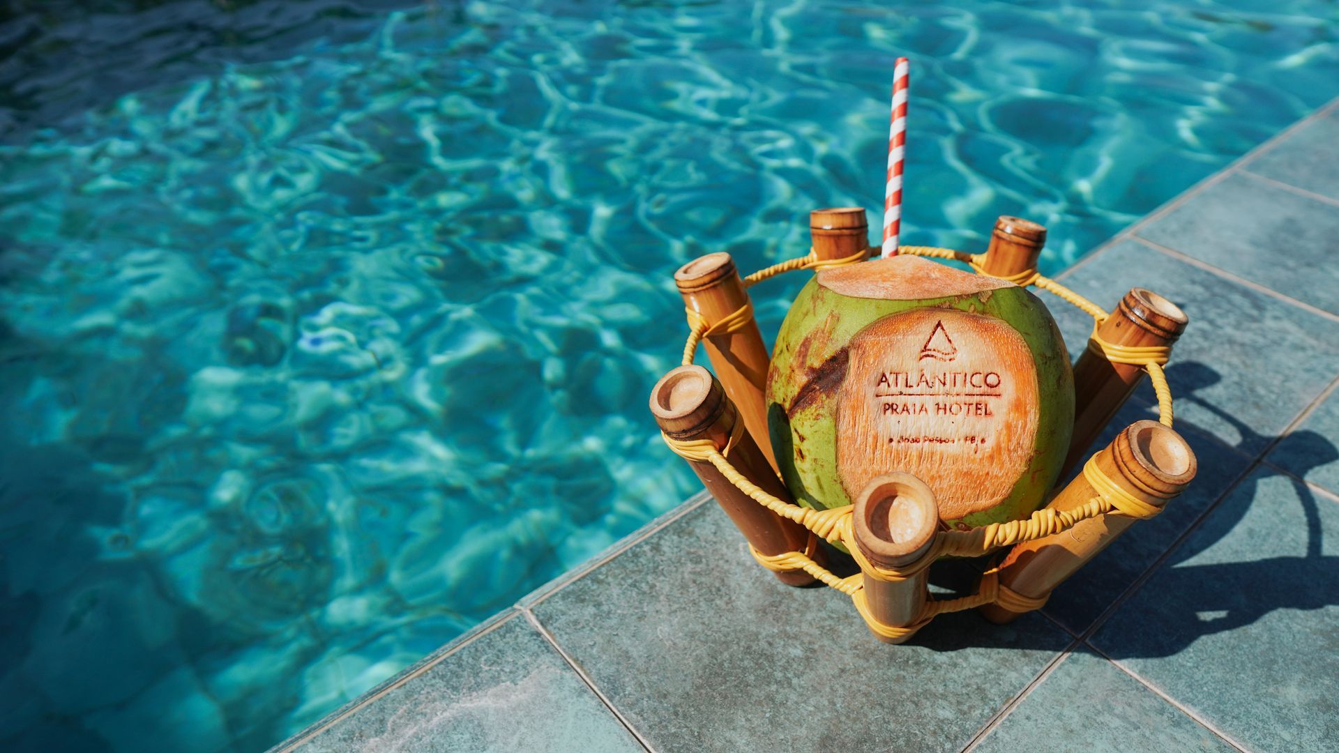 Um coco com a marca estampada e um canudo, acomodado em um suporte de bambu na borda de uma piscina azul vibrante.