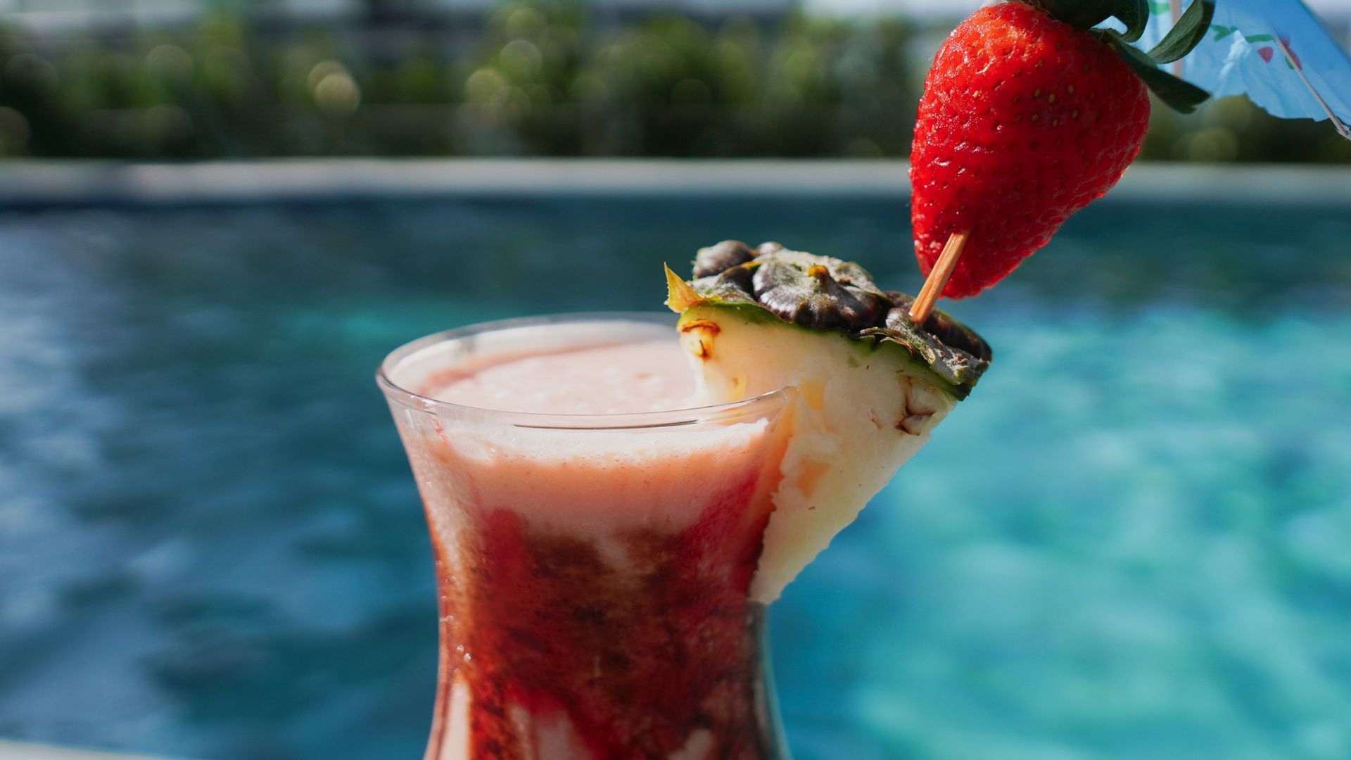 Um coquetel de frutas com decoração de morango e abacaxi, tendo como pano de fundo uma piscina desfocada.