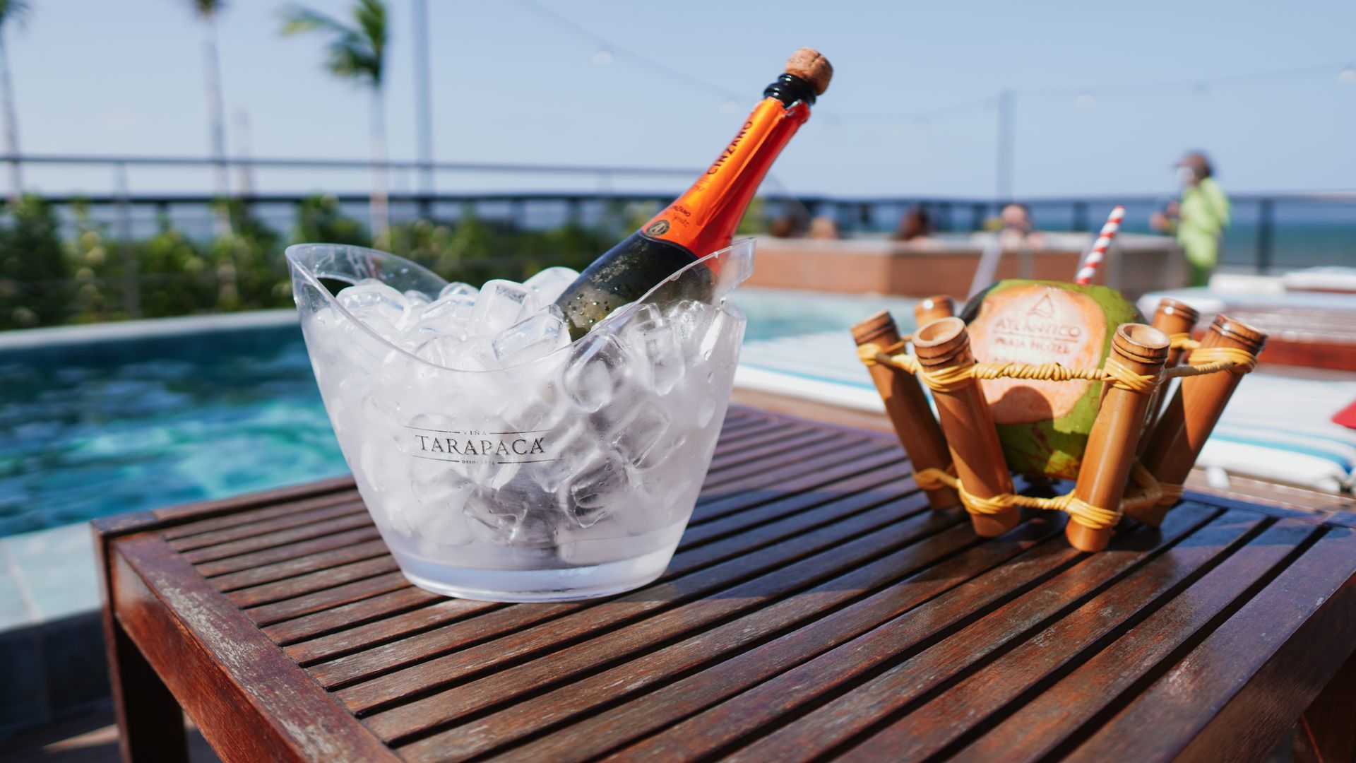 Uma garrafa de champanhe em um balde de gelo e uma bebida de coco sobre uma mesa de madeira ao lado de uma piscina sob um céu claro.