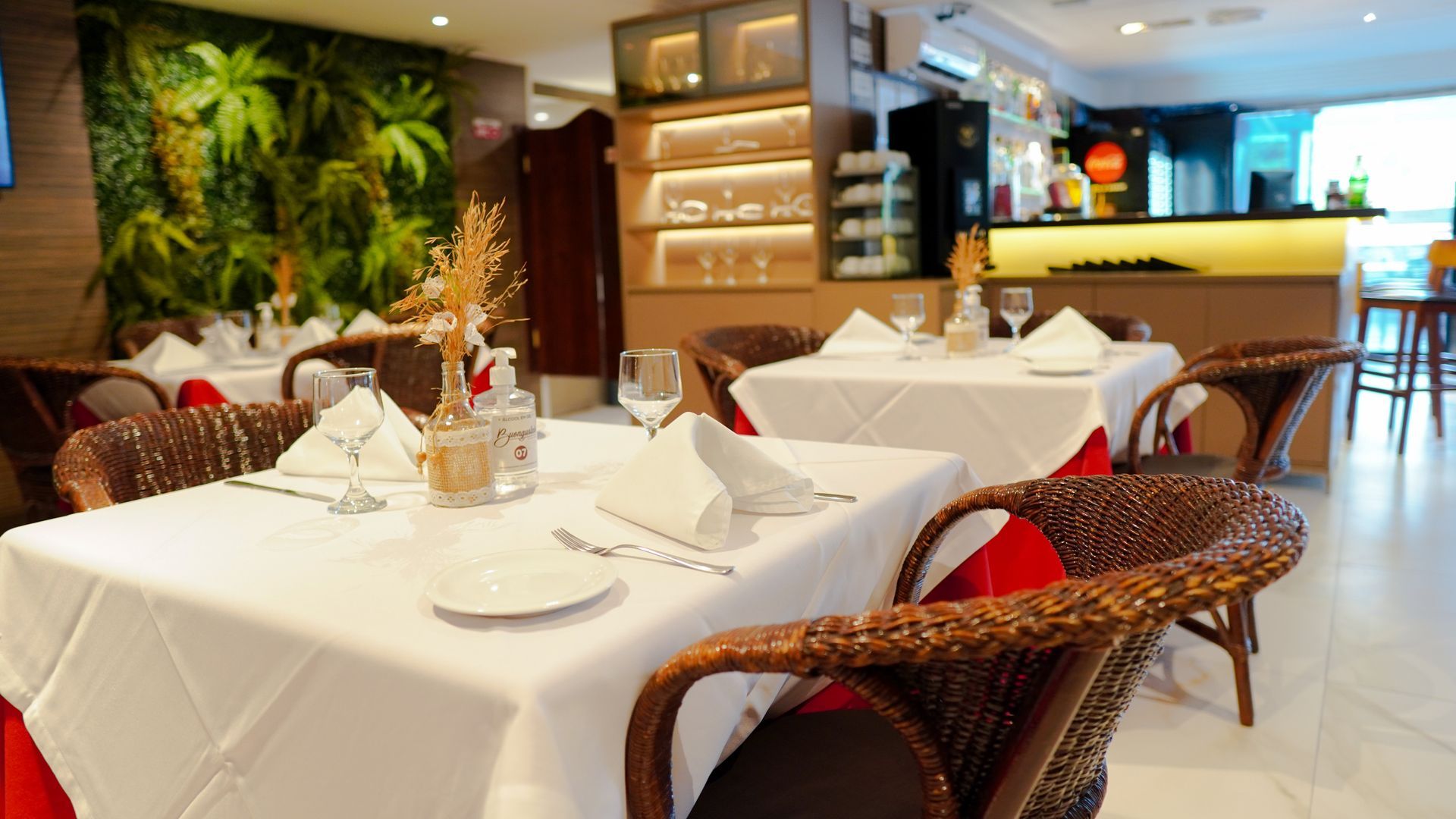 Uma área de jantar de restaurante bem iluminada, com mesas postas com toalhas de mesa brancas, guardanapos e cadeiras de vime marrom.