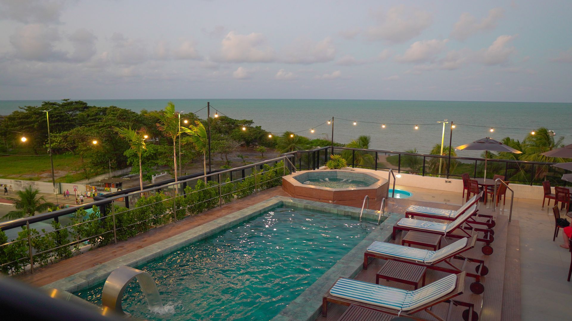 Vista panorâmica de uma piscina retangular e uma banheira de hidromassagem em um deck com vista para o oceano ao entardecer.