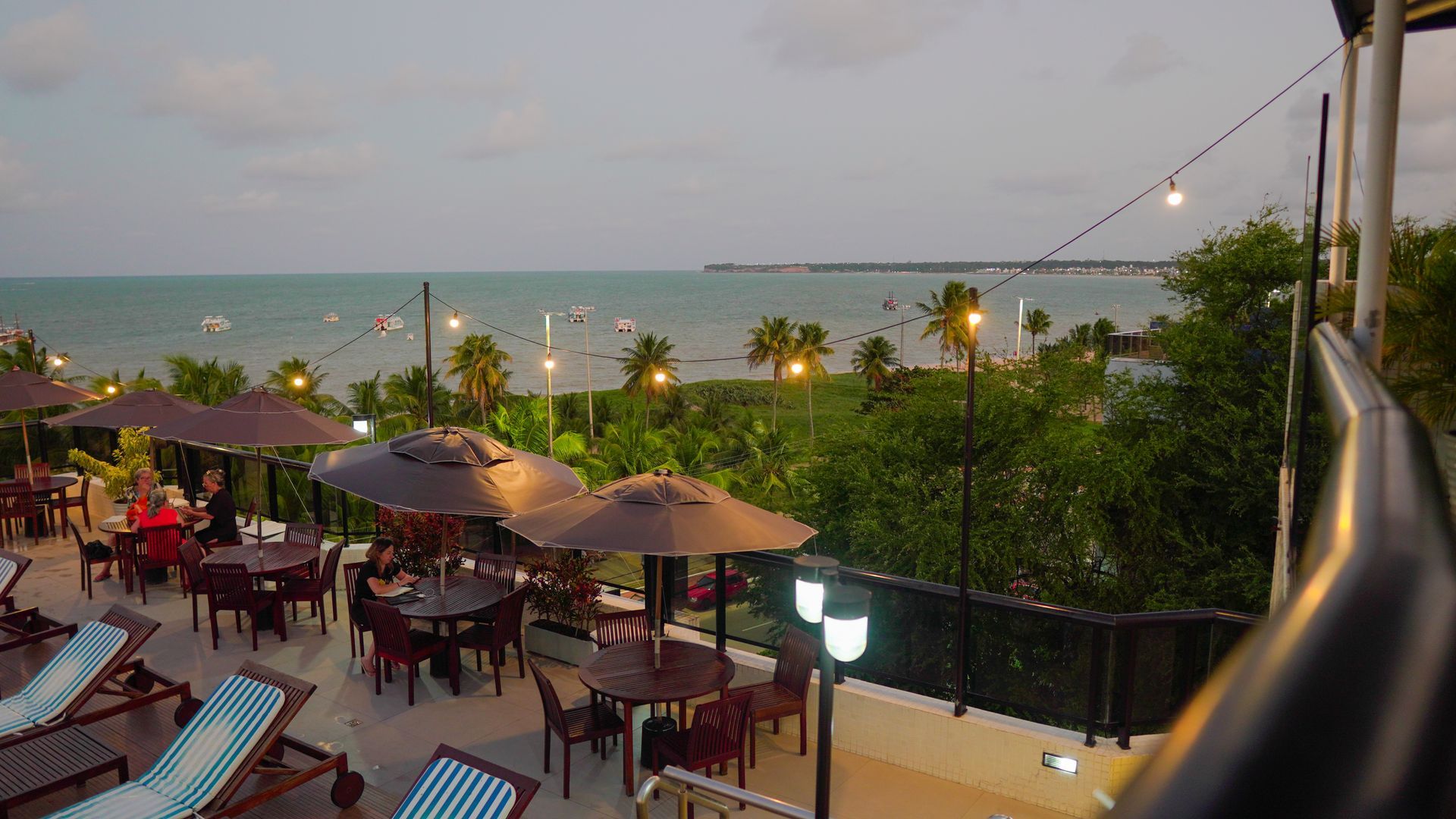 Um terraço elevado ao ar livre, decorado com luzes de corda e mesas de jantar, oferece vista para o oceano calmo ao entardecer.