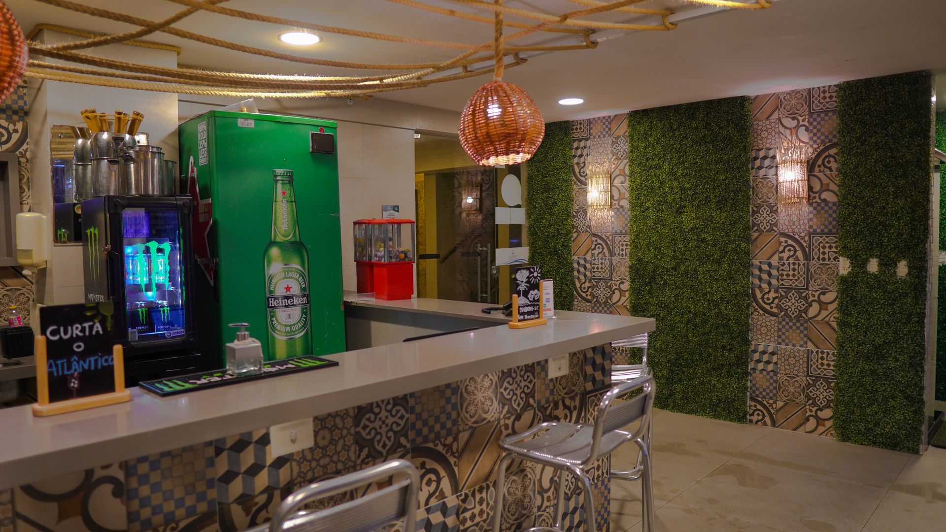 Um balcão de bar com refrigerador de cerveja, banquetas, detalhes em azulejos estampados e vegetação vertical na parede sob iluminação de vime.