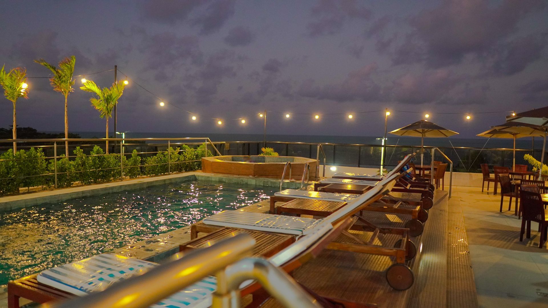 Um terraço com piscina ao entardecer, com espreguiçadeiras, mesinhas e luzes de corda com vista para o oceano.