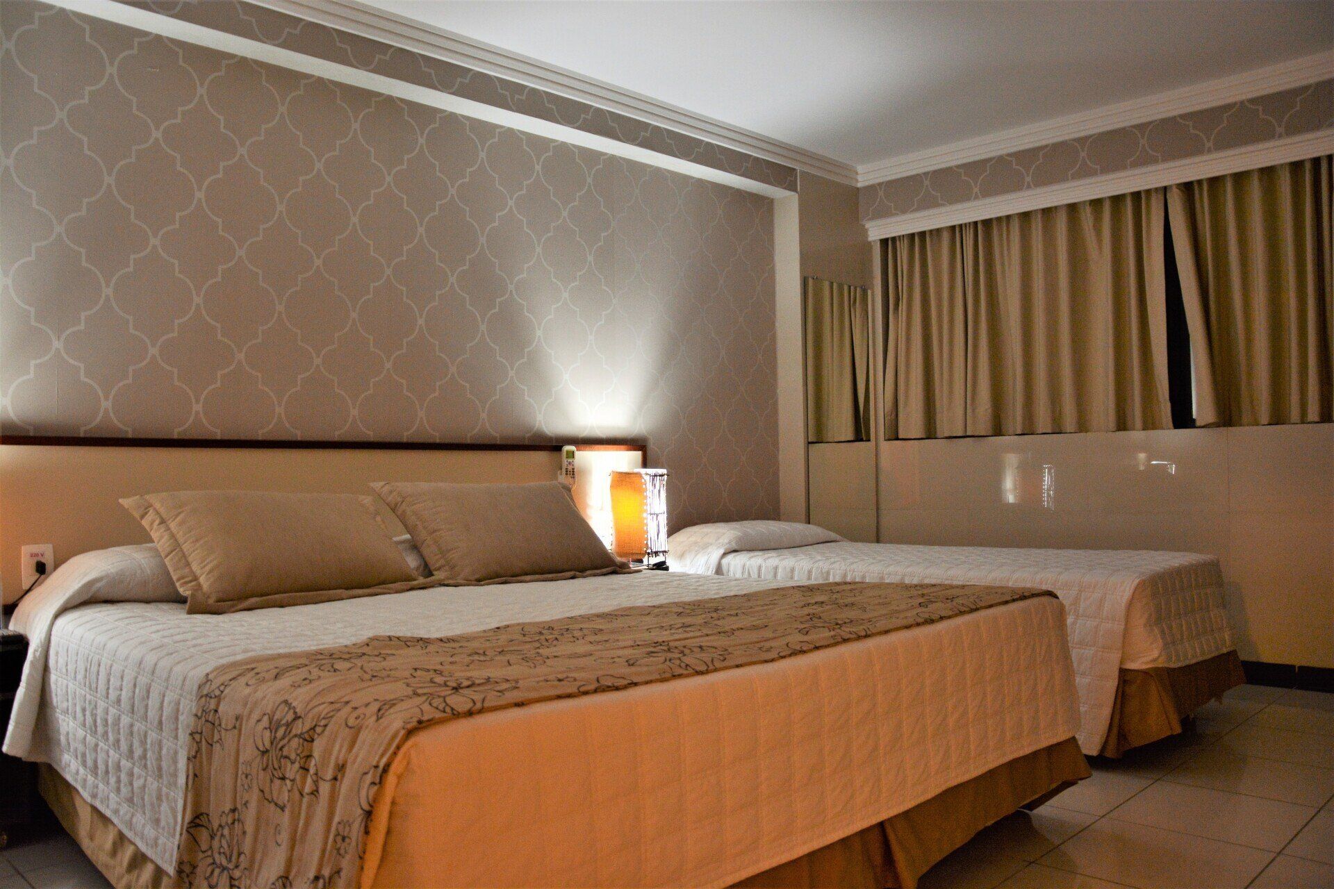 Um quarto de hotel com uma cama grande em primeiro plano e uma cama de solteiro menor ao fundo, ambas com roupa de cama bege estampada.