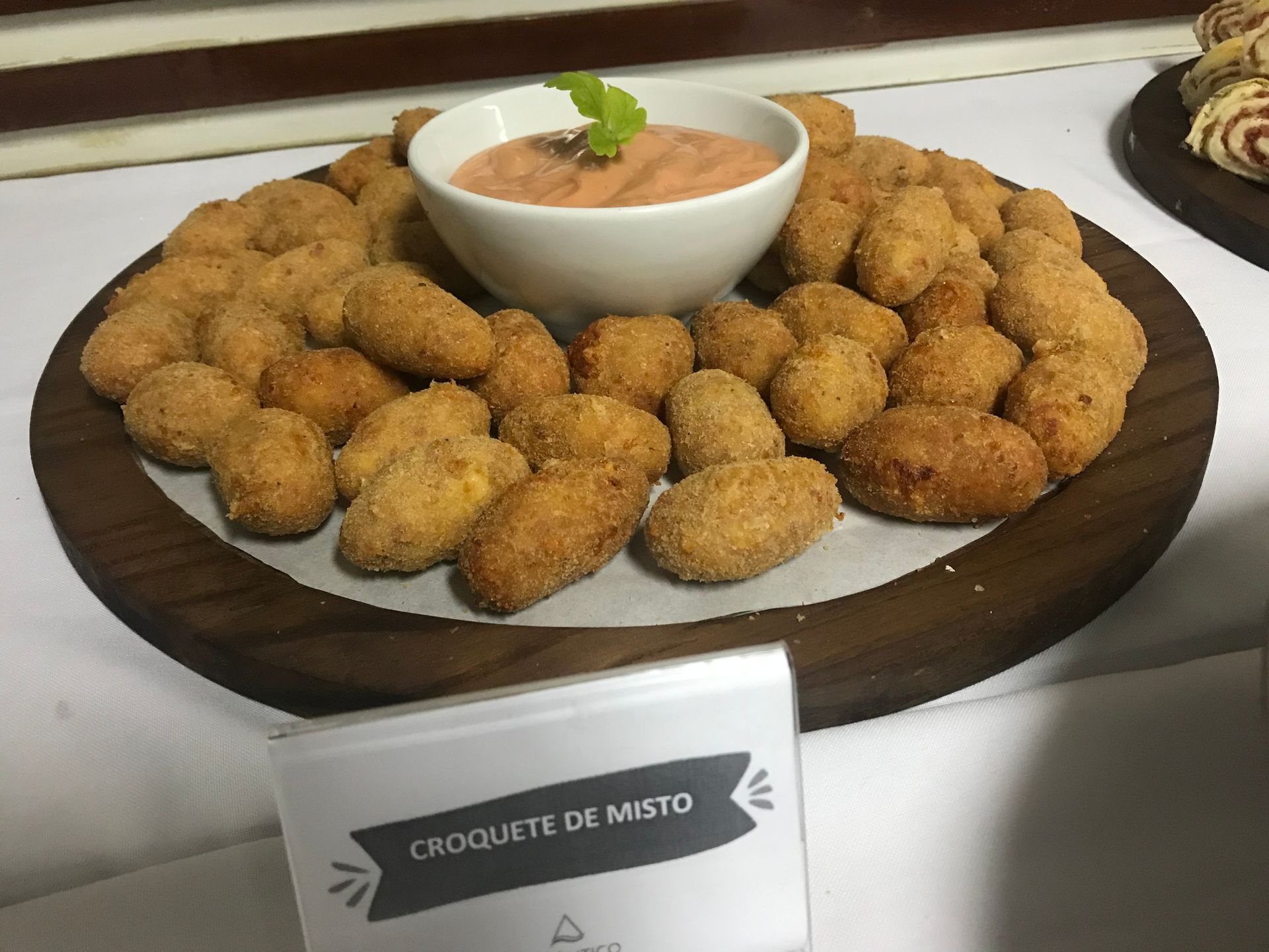 Uma travessa de madeira contém croquetes fritos dourados, dispostos ao redor de uma tigela central com molho rosa e uma guarnição.