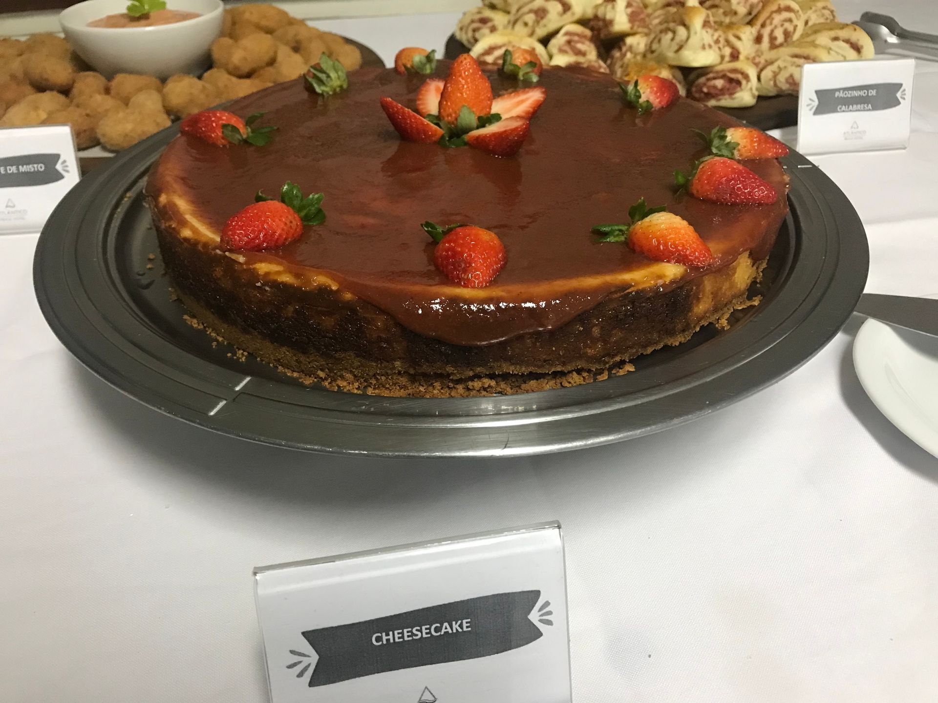 Uma grande cheesecake coberta com uma cobertura brilhante e morangos frescos, disposta numa travessa sobre a mesa com outros petiscos.
