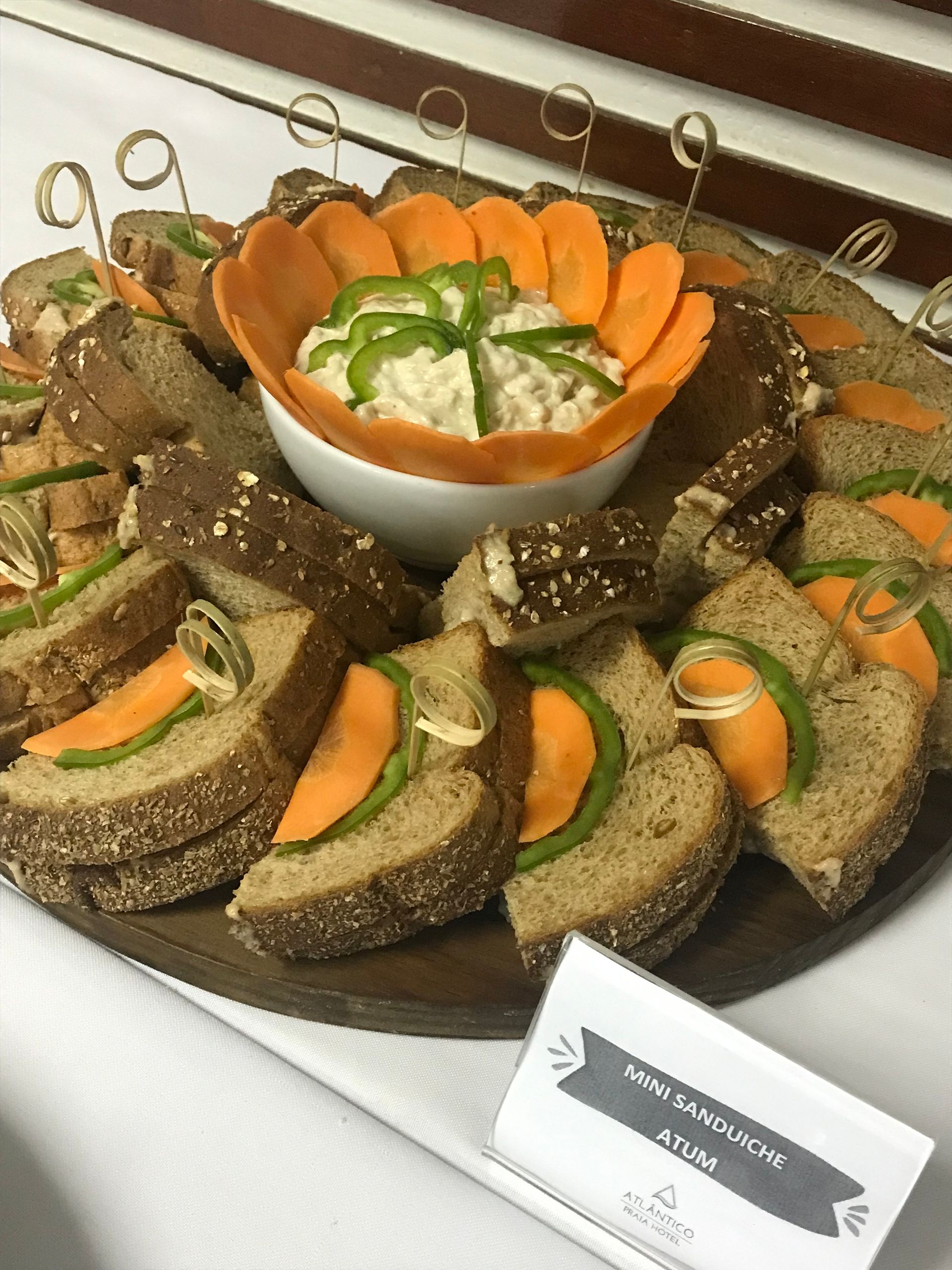 Uma travessa com mini sanduíches abertos de pão de centeio, cobertos com fatias de cenoura e pimentão, e um recipiente central para molho.
