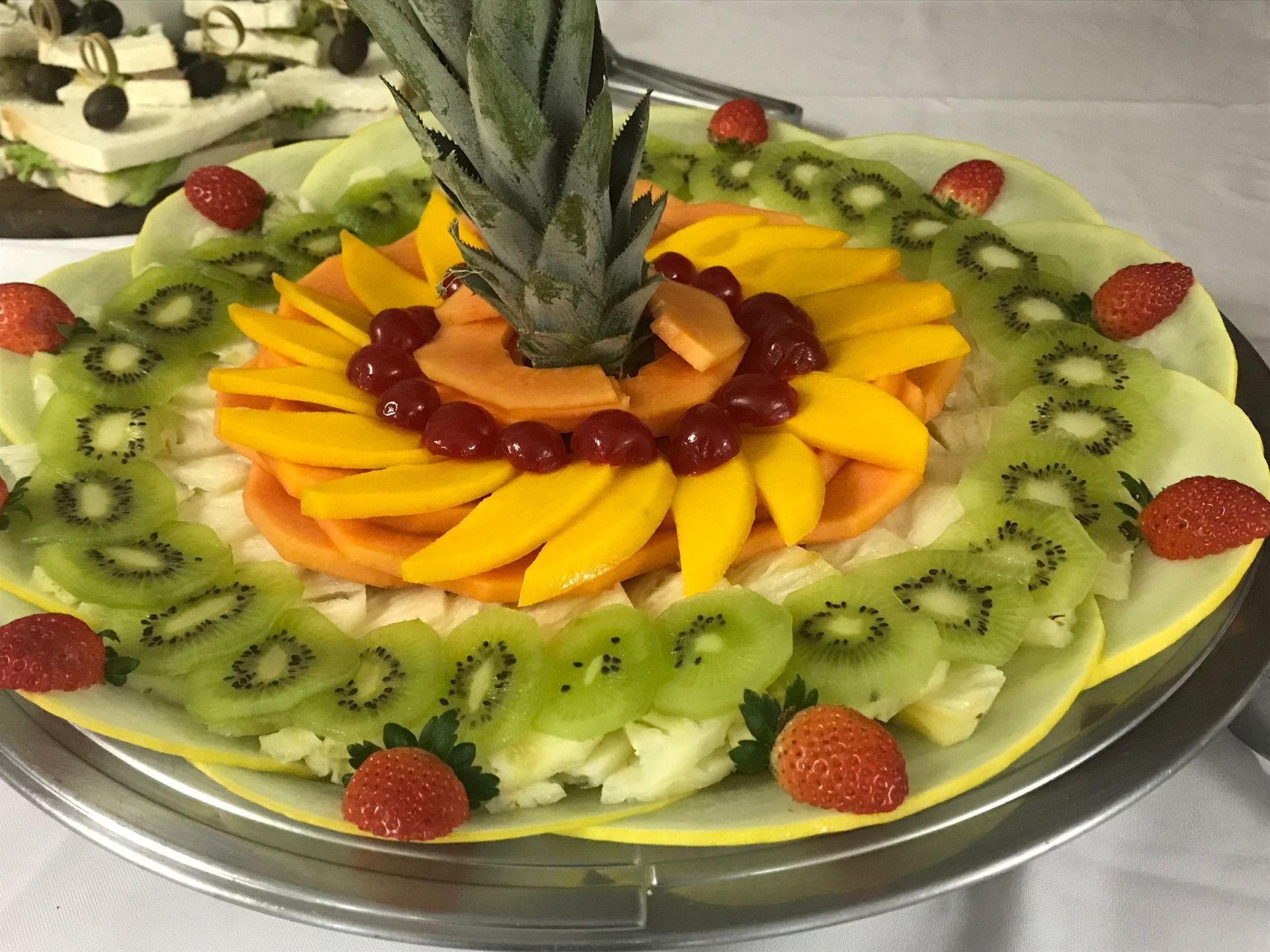 Uma travessa decorativa de frutas com abacaxi, kiwi fatiado, manga, uvas e morangos dispostos em uma bandeja circular.