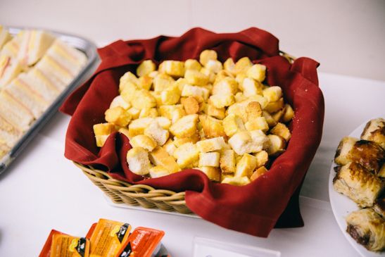 Uma cesta de croutons dourados forrada com um guardanapo cor de vinho, colocada sobre uma mesa com sanduíches e doces.