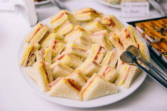 Um prato branco repleto de sanduíches triangulares de presunto e queijo, com uma pinça de metal para servir ao lado.