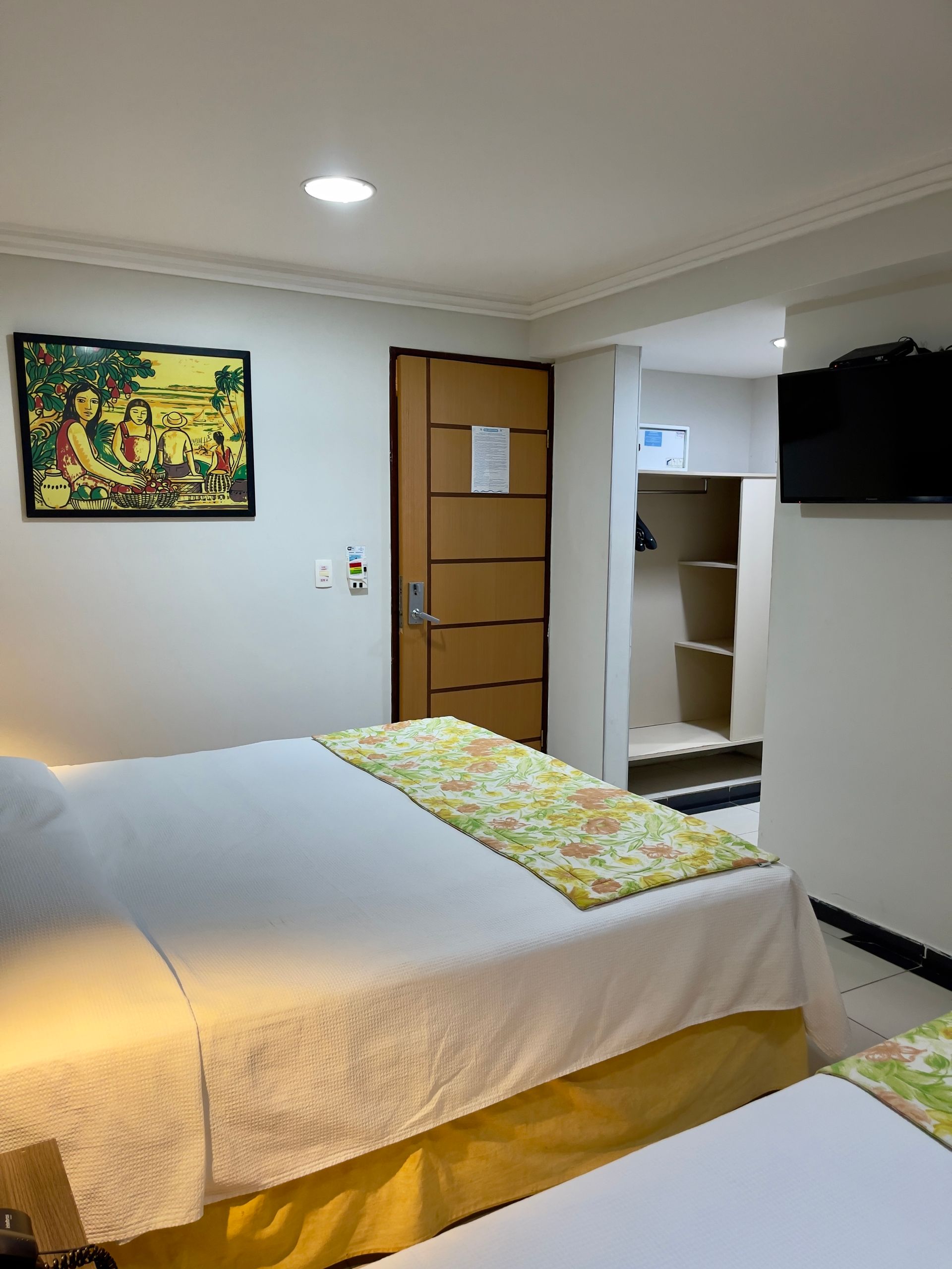 Um quarto de hotel arrumado com uma cama de lençóis amarelos, um quadro na parede, uma porta de madeira e uma televisão na parede.
