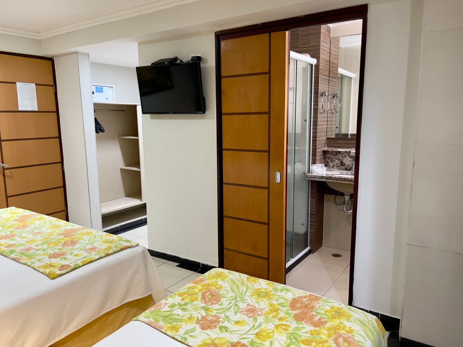 Um quarto de hotel com duas camas com peseiras florais, uma TV fixada na parede, um armário aberto e uma porta de banheiro adjacente.
