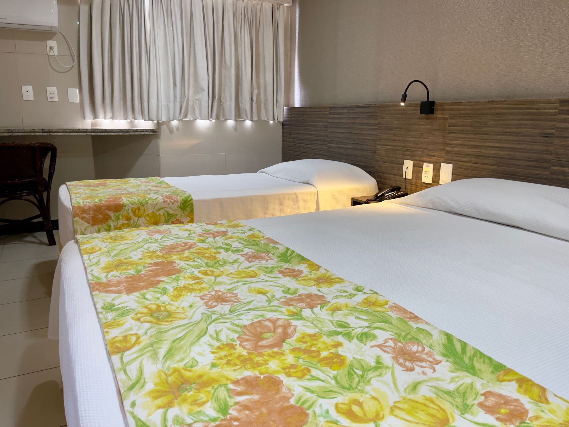 Um quarto de hotel com duas camas brancas com caminhos de cama florais, cabeceira de madeira e cortinas brancas.