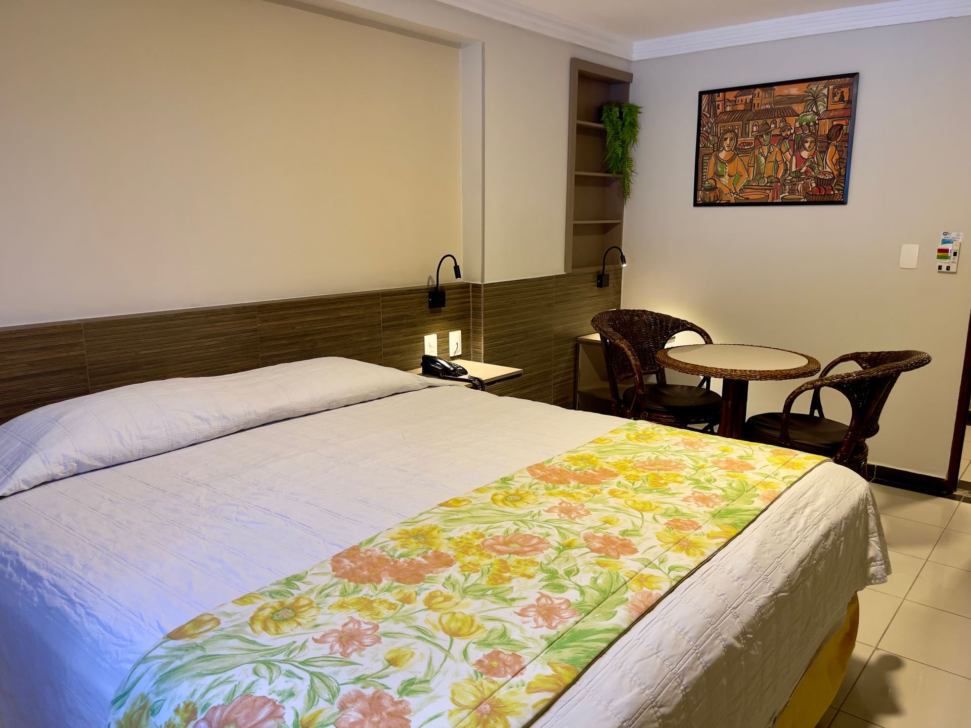 Um quarto de hotel com uma cama com um caminho de mesa floral, uma pequena mesa, duas cadeiras e um quadro na parede.