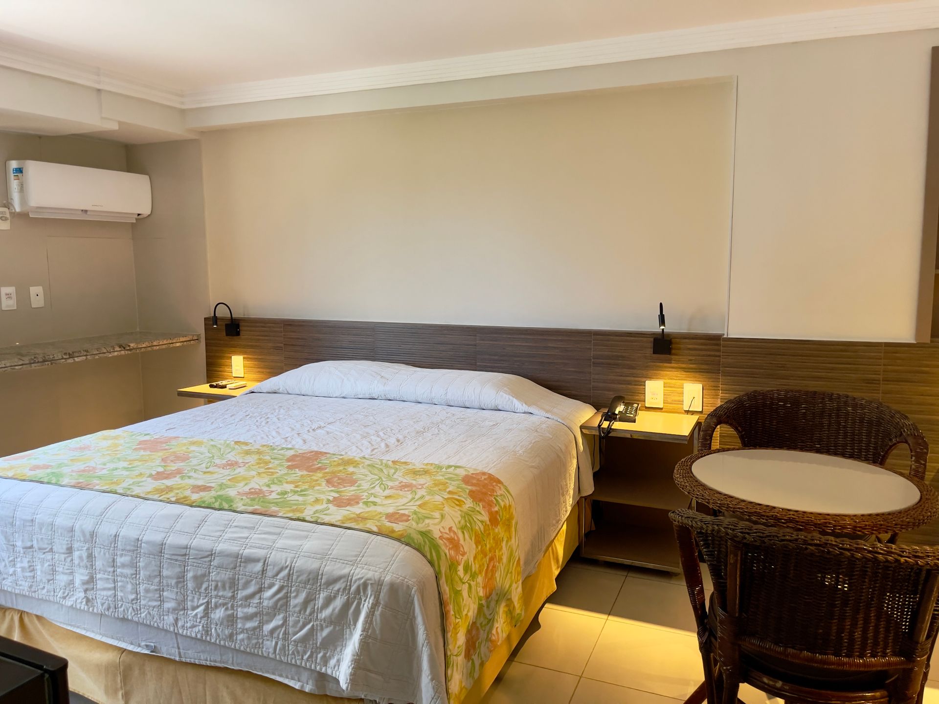 Um quarto de hotel com uma cama queen-size com um caminho de mesa floral, paredes claras, cabeceira de madeira e uma pequena mesa e cadeiras de vime.