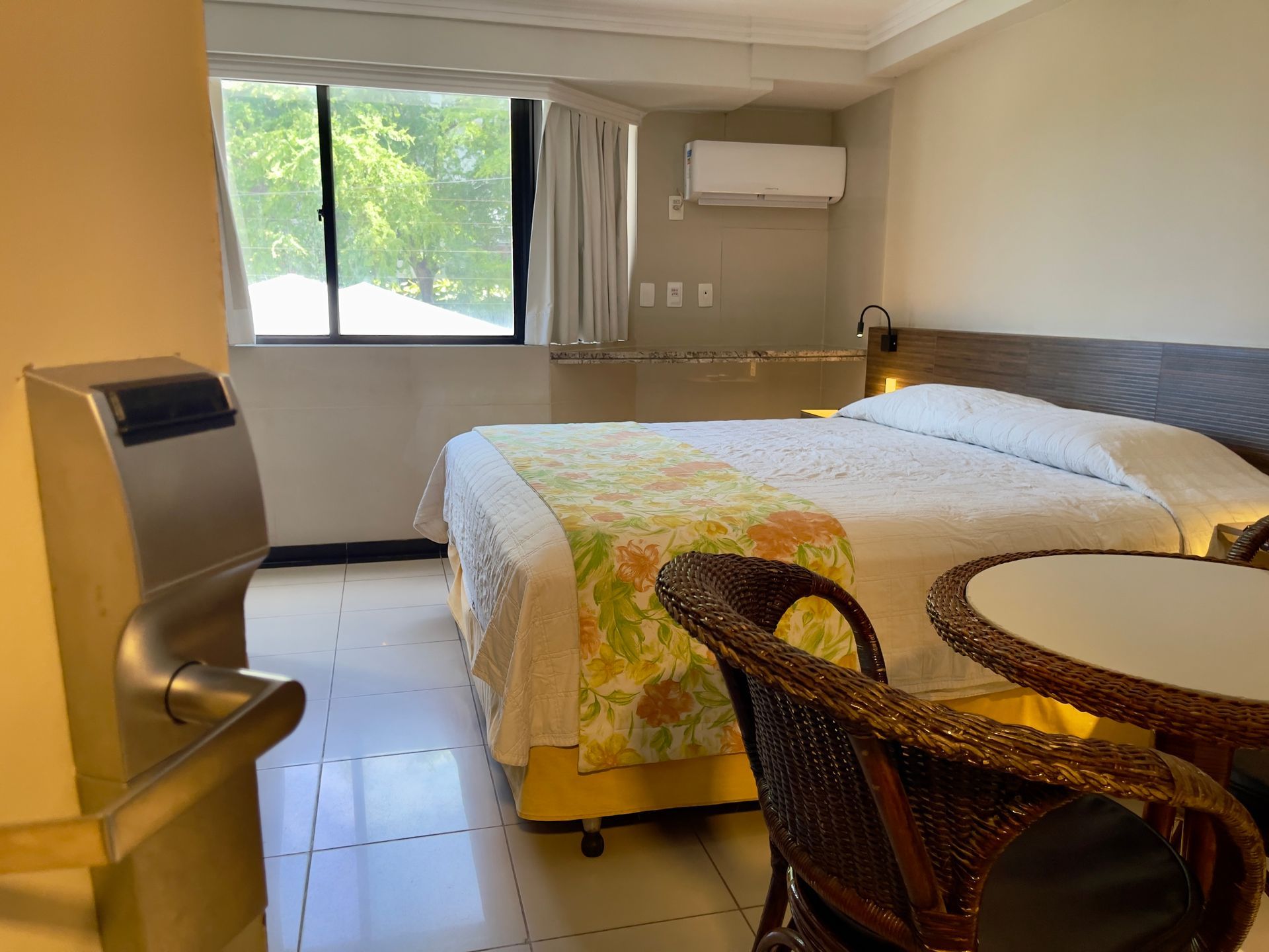 Um quarto de hotel com uma cama queen-size com um caminho de mesa floral, uma pequena mesa com cadeiras de vime e uma janela com cortinas brancas.