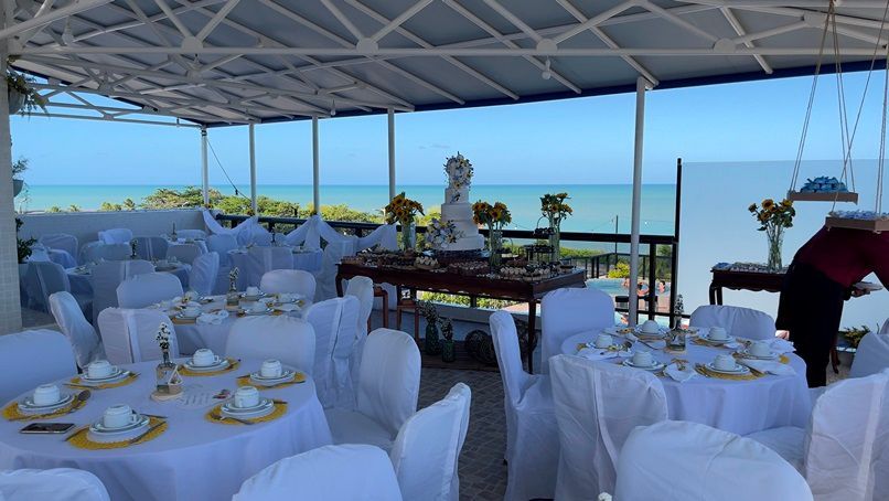 Um espaço para eventos ao ar livre com mesas e cadeiras cobertas com toalhas brancas e um bolo como peça central, tendo o oceano como pano de fundo.