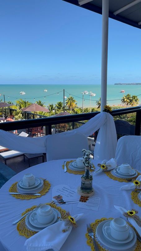 Uma mesa posta com toalhas brancas e detalhes de girassóis, com vista para o litoral, o oceano azul e barcos ao longe.