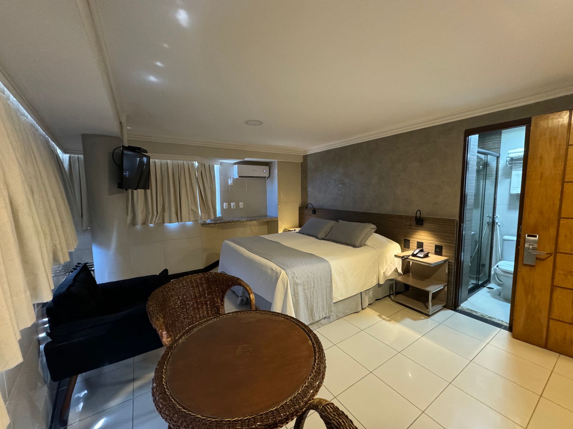 Um quarto de hotel com uma cama queen-size, piso de azulejo claro, uma pequena mesa de vime com duas cadeiras e um banheiro privativo.