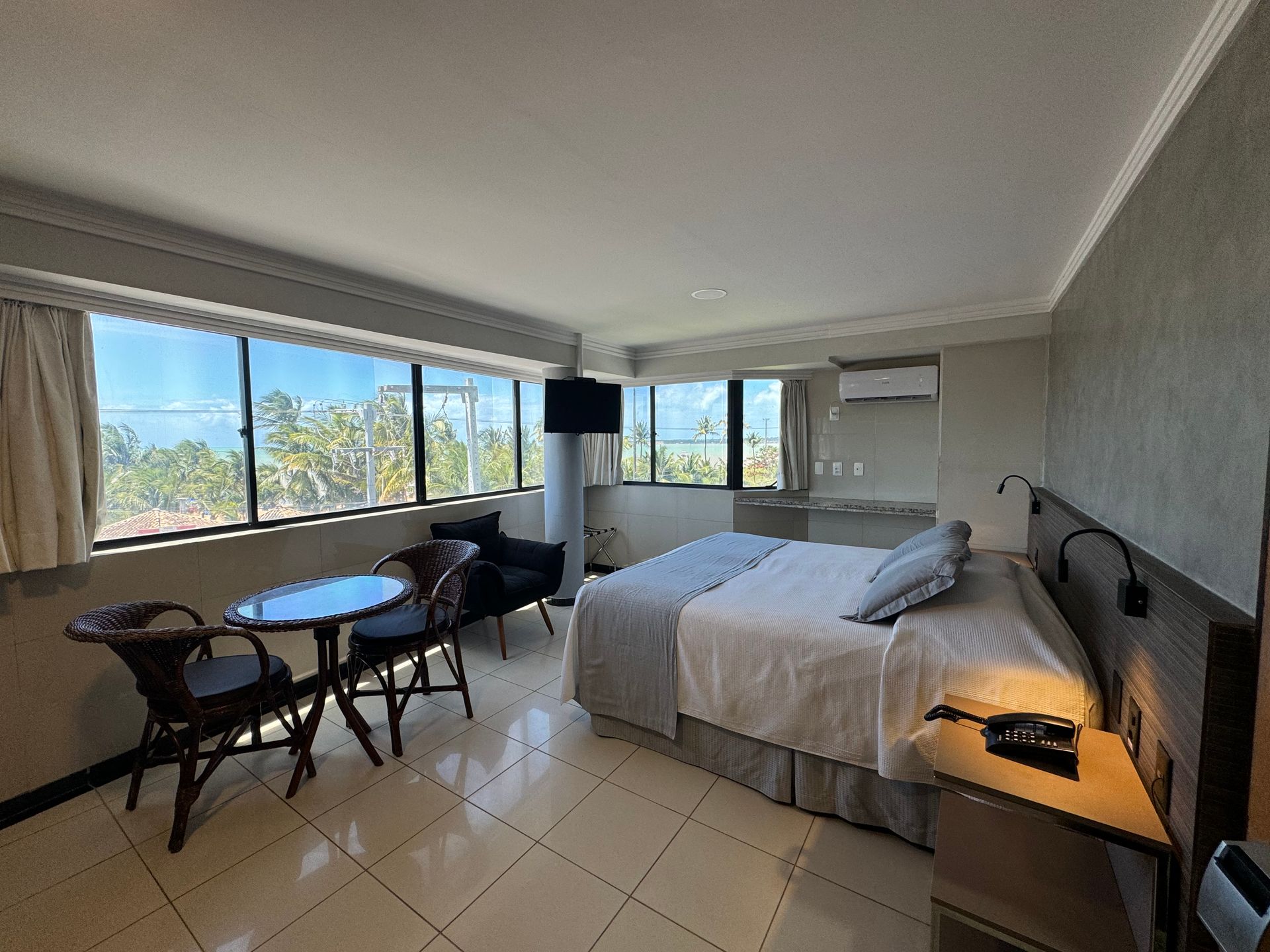 Um quarto de hotel com uma cama queen-size, uma pequena mesa de jantar com duas cadeiras, uma poltrona e janelas do chão ao teto.