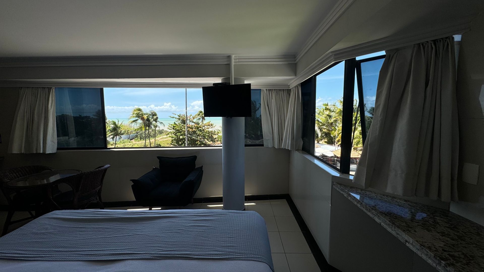 Um quarto de hotel com cama, poltrona e janelas do chão ao teto com vista para uma paisagem tropical ensolarada.