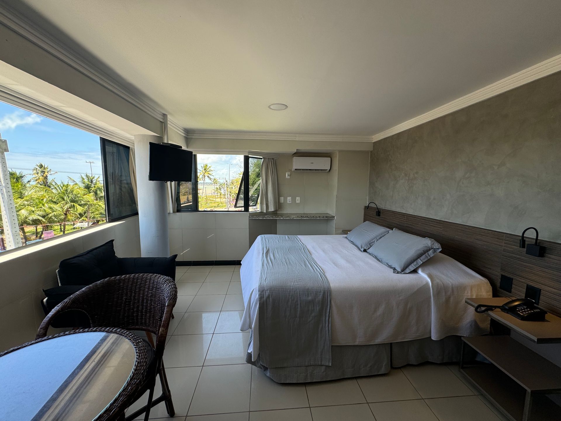 Um quarto de hotel moderno e bem iluminado, com uma cama grande, varanda com vista para palmeiras e uma pequena área de estar.