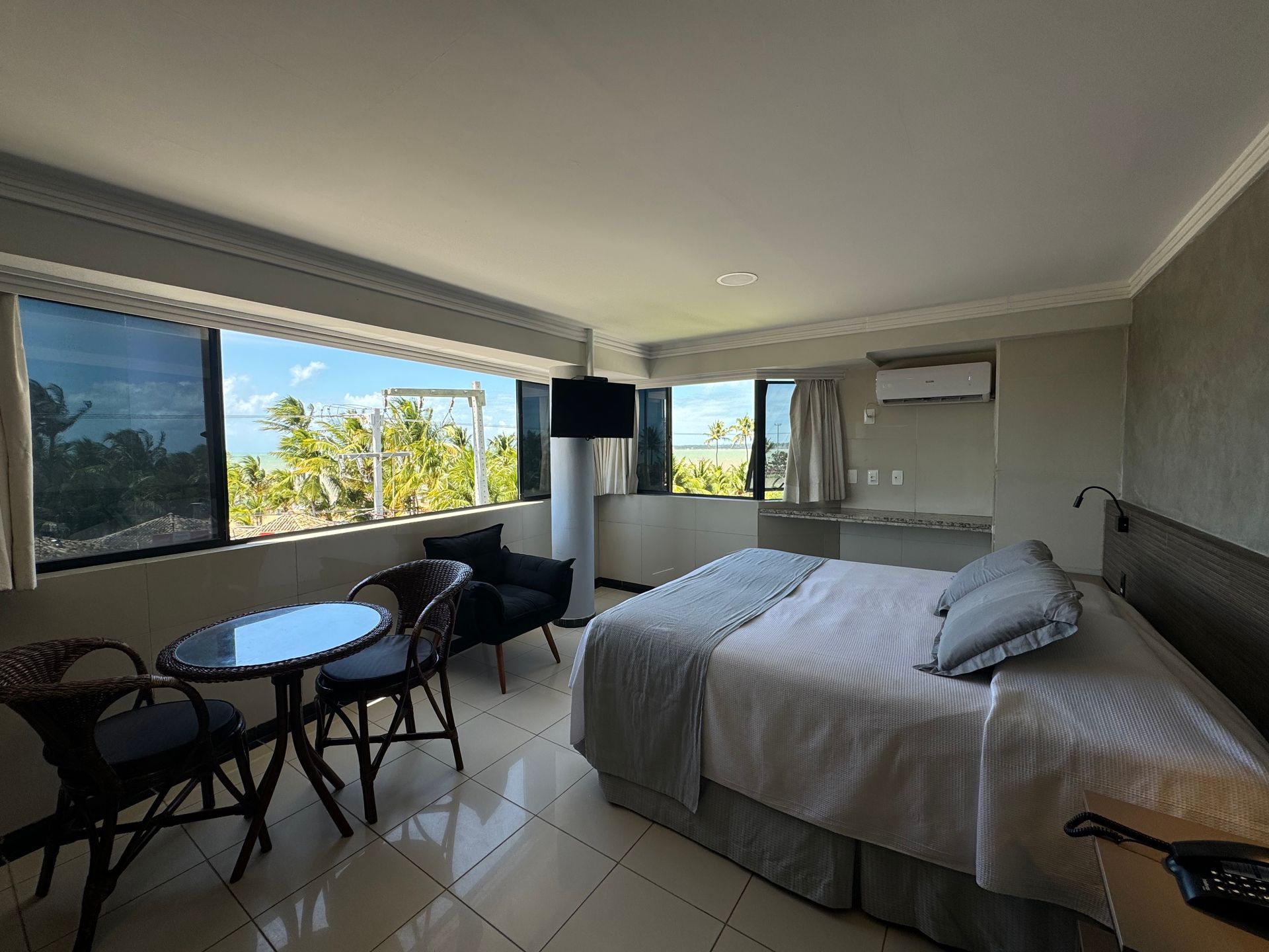 Um quarto de hotel moderno com uma cama grande, área de estar e uma janela panorâmica com vista para uma paisagem ensolarada de praia.